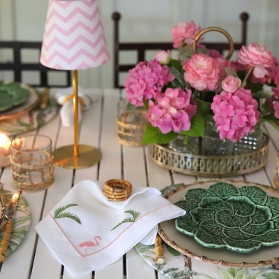 Mesa posta com toalha branca, prato verde decorativo, flores rosas e abajur com sombra rosa de padrão zigzag