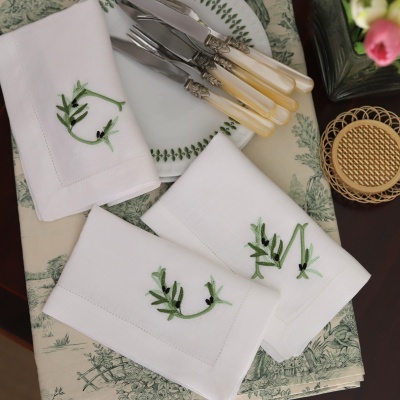 Guardanapos de tecido branco com bordado verde, facas com cabos perolados, prato branco com motivo de folhas verdes sobre toalha estampada