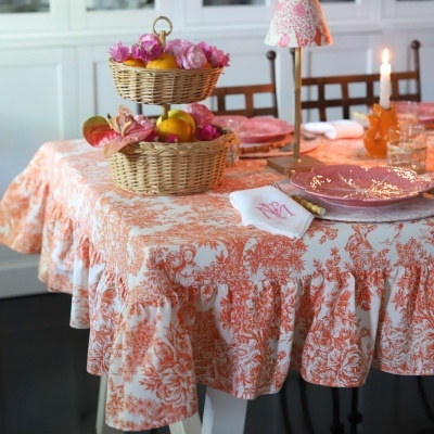 Mesa posta com toalha laranja e branca, cesta de vime com flores e frutas, pratos rosas em forma de folha, guardanapo branco com monograma.