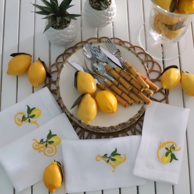 Talheres com cabos de madeira, limões artificiais, guardanapos bordados com limões e plantas decorativas numa mesa branca.