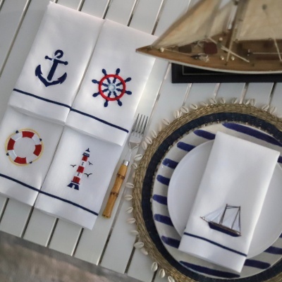 Quatro guardanapos brancos bordados com motivos náuticos coloridos sobre mesa com prato e vela bordada