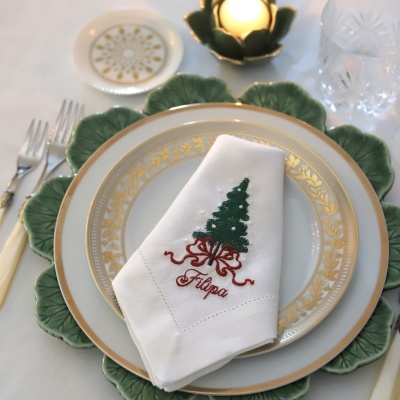 Lugar de mesa com prato branco e dourado e guardanapo branco bordado com árvore de Natal e nome 'Filipa'