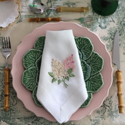 Conjunto de louça com prato rosa, prato verde em padrão de folhas, guardanapo branco bordado, talheres com cabos em bambu e toalha verde estampada.