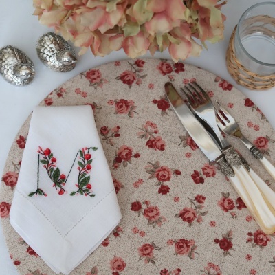 Talheres com cabo creme, guardanapo bordado com letra M floral, suporte de tecido com rosas vermelhas, saleiros prateados e copo com suporte de vime