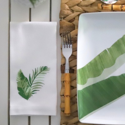 Guardanapo branco com folhas verdes, prato quadrado branco com folhas verdes e garfo de cabo de madeira sobre base de fibra trançada