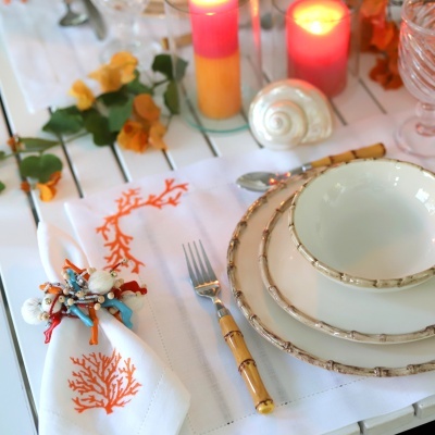 Jogos de jantar em cerâmica creme com bordas castanhas num arranjo decorativo com vela, flor seca e guardanapo com coral laranja.