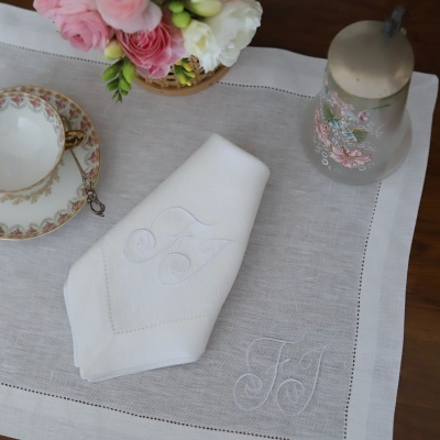 Toalha e guardanapo de linho branco com bordado FJ e chávena, flores, recipiente decorado sobre mesa de madeira.