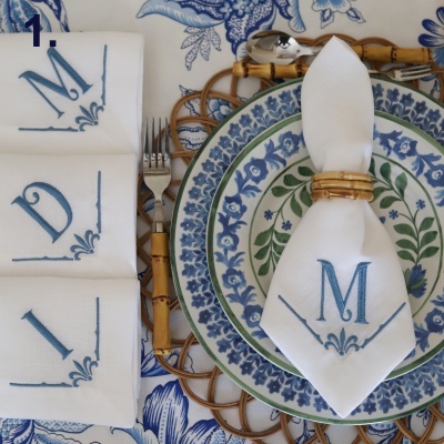 Jogo de guardanapos bordados com letras D, M e L ao lado de prato decorado e guardanapo no centro com anel