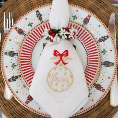 Guardanapo branco bordado com nome Sara segurado por argola decorativa em prato com padrão de soldadinhos de chumbo natalícios.