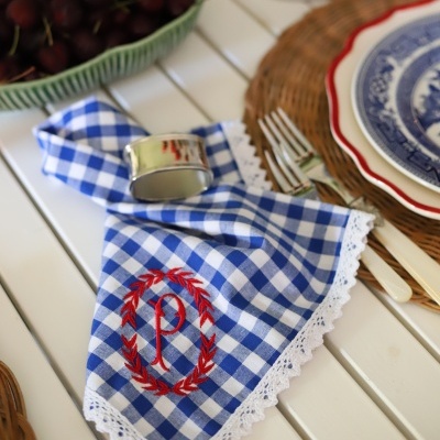 Guardanapo quadriculado azul e branco com bordado vermelho e renda branca, anel de guardanapo prateado, talheres e pratos sobre mesa branca