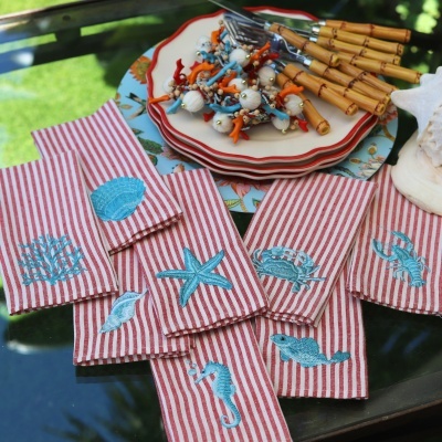Toalhas riscas vermelhas e brancas com bordados azuis de temas marinhos numa mesa de vidro com pratos e talheres.