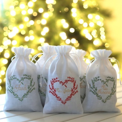 Cinco sacos brancos de tecido com corações estampados e texto dourado, luzes de natal ao fundo