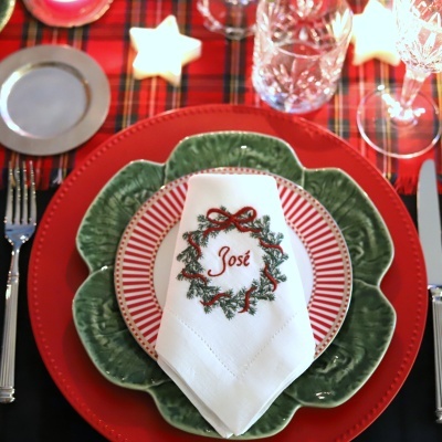 Mesa posta com pratos vermelho e verde, guardanapo branco bordado com 