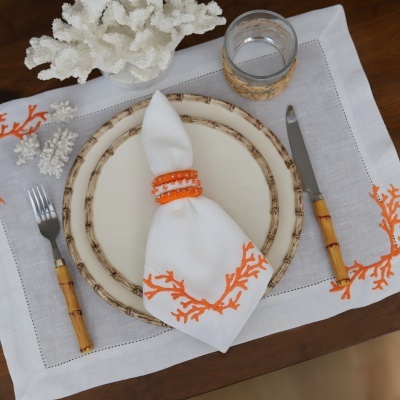 Mesa posta com guardanapo branco bordado, pratos creme, talheres com cabo em bambu e copo com suporte rústico.