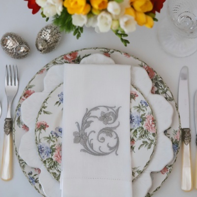 Mesa posta com pratos florais, talheres com cabos bege e guardanapo branco bordado