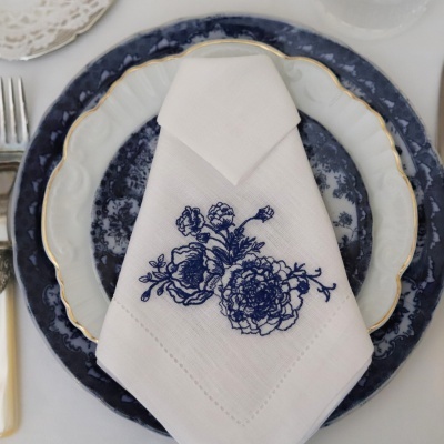 Pratos de porcelana com borda azul escura e dourada com guardanapo branco bordado a azul com flores.