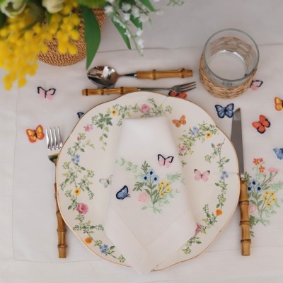 Conjunto de loiça floral com talheres de cabo madeira e copo com suporte rústico sobre toalha branca decorada com borboletas coloridas.