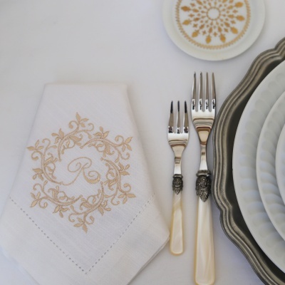 Toalha branca com bordado dourado, garfos com cabos brancos e pratos brancos e cinza