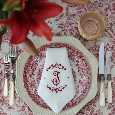 Mesa posta com louça creme e vermelha, guardanapo bordado, copo com suporte de palhinha e flores vermelhas.