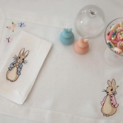 Guardanapo e toalha branca de linho bordados com coelhos, velas azul e pêssego, cloche de vidro e taça com amêndoas coloridas