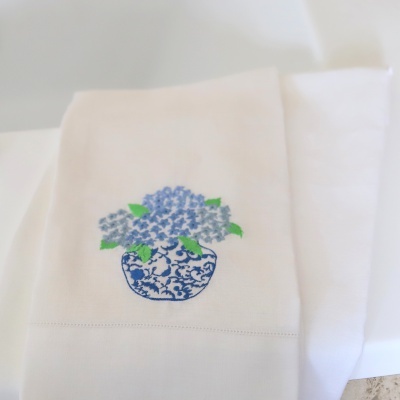 Toalha branca com bordado de vaso azul e flores lilás.