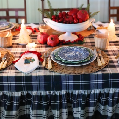 Mesa posta com toalha xadrez azul e branca, pratos, guardanapo com ursinho bordado e velas decorativas de Natal acesas.