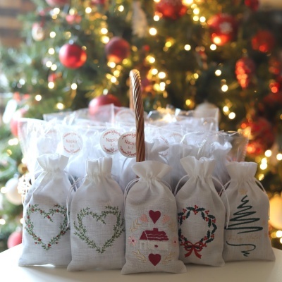 Cinco sacos de tecido branco com bordados natalícios em frente a cesta com mais sacos 'Napkin & Co' e árvore de Natal iluminada ao fundo