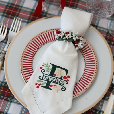Guardanapo branco bordado com nome Francisca e decoração floral em pratos com padrão riscas vermelhas e douradas sobre toalha xadrez