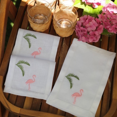 Guardanapos de tecido com bordados de flamingos e folhas, copos com suporte de vime numa mesa de madeira com flores cor-de-rosa.