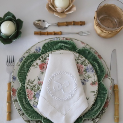 Mesa posta com pratos florais, prato verde texturizado, talheres com cabo de bambu, guardanapo branco bordado, copo com suporte em vime e velas em suportes florais.