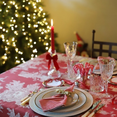 Mesa de jantar com decoração natalícia, toalha vermelha e branca, pratos, talheres, guardanapo, castiçal com vela vermelha e copos de cristal.