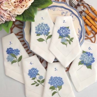 Seis guardanapos brancos bordados com flores azuis, dispostos sobre prato branco com azul e ao lado de talheres e flores