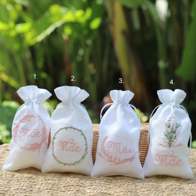 Quatro saquinhos brancos bordados com a palavra 'Mãe' e elementos florais, em fundo natural