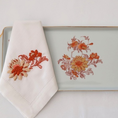 Guardanapo branco bordado com flores laranja ao lado de prato cinza com padrão floral