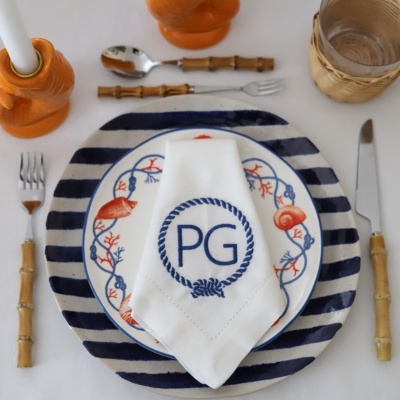 Mesa posta com prato decorado com corais e conchas, talheres com cabos de bambu, guardanapo branco bordado com as letras PG, castiçal e jarro laranja, copo com suporte de vime.