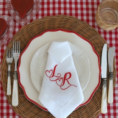 Conjunto de loiça com prato grande com borda vermelha e prato pequeno branco, guardanapo branco bordado com letras vermelhas L e R, talheres com cabos brancos e base de vime sobre toalha xadrez vermelha e branca