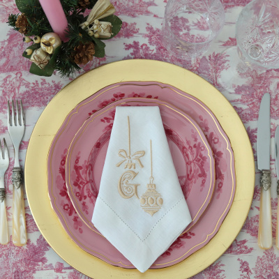 Mesa posta com pratos rosa e dourados, copos de vidro e guardanapo branco bordado