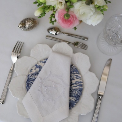 Conjunto de louça branco e azul com guardanapo bordado, talheres e arranjo floral numa mesa branca