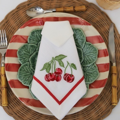 Conjunto de loiça com prato vermelho e branco às riscas, prato verde com folhas, guardanapo com cerejas bordadas e talheres com cabo castanho