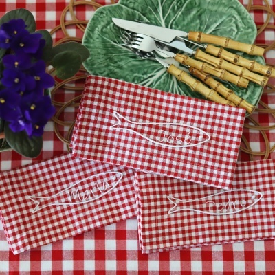 Guardanapos às quadrículas vermelhas e brancas com nomes bordados, talheres com cabo de bambu, prato verde e planta com flores roxas numa mesa com toalha xadrez