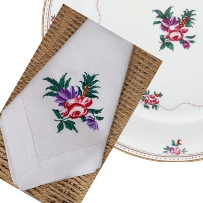 Guardanapo branco bordado com flores coloridas ao lado de prato branco com padrão floral similar