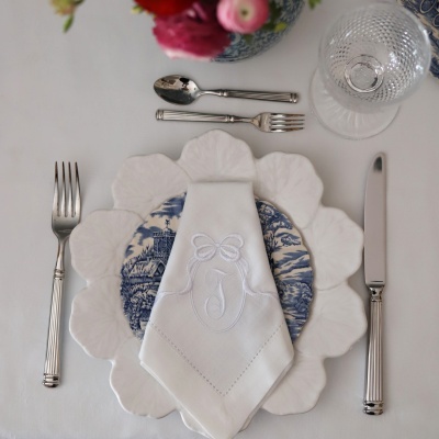 Conjunto de mesa com pratos, guardanapo branco monogramado, talheres de metal e copo de vidro texturizado