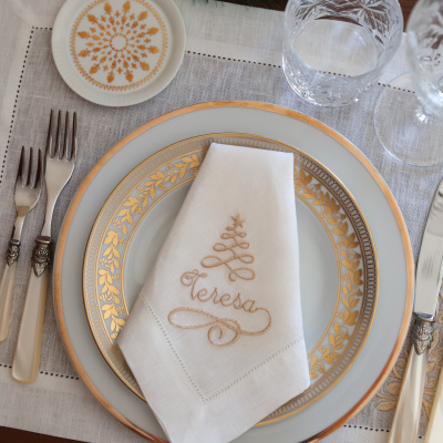 Conjunto de louça branca com padrão dourado, guardanapo branco bordado, garfos com cabo creme e copo de vidro transparente sobre toalha branca