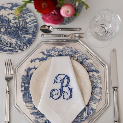 Mesa posta com pratos em padrão azul e branco, guardanapo branco bordado com letra B, talheres prateados e copo de vidro. Arranjo floral no centro.