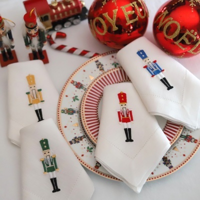 Guardanapos brancos com bordados de soldadinhos de chumbo em vários tons, prato decorado com soldadinhos, bolas vermelhas de Natal e brinquedos na mesa.