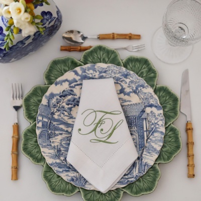 Conjunto de loiça e talheres com prato verde e azul, guardanapo branco bordado, talheres com cabos de bambu e copo de vidro.