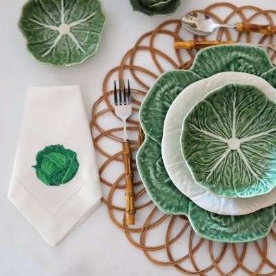 Pratos verdes com padrão de folhas, guardanapo branco com bordado de alface, talheres e sousplat trançado.