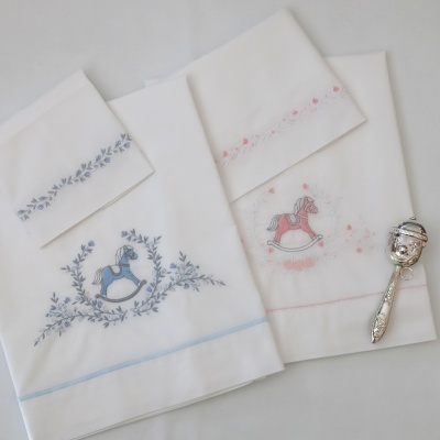 Lençóis de berço brancos com bordados de cavalinhos azul e rosa e chocalho prateado