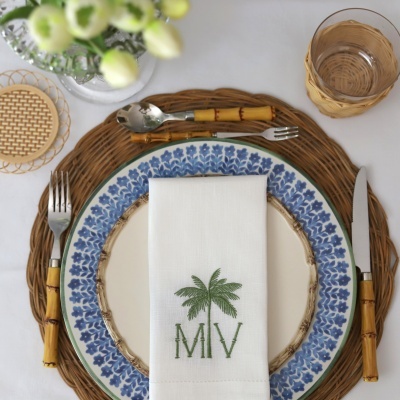 Conjunto de louça azul e bege com guardanapo branco bordado, talheres com cabos de bambu, base de rattan e copo com suporte de rattan