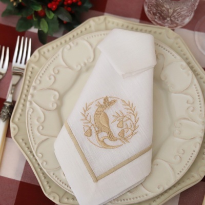 Guardanapo branco bordado com pássaro dourado sobre prato decorado creme em mesa xadrez vermelha e branca